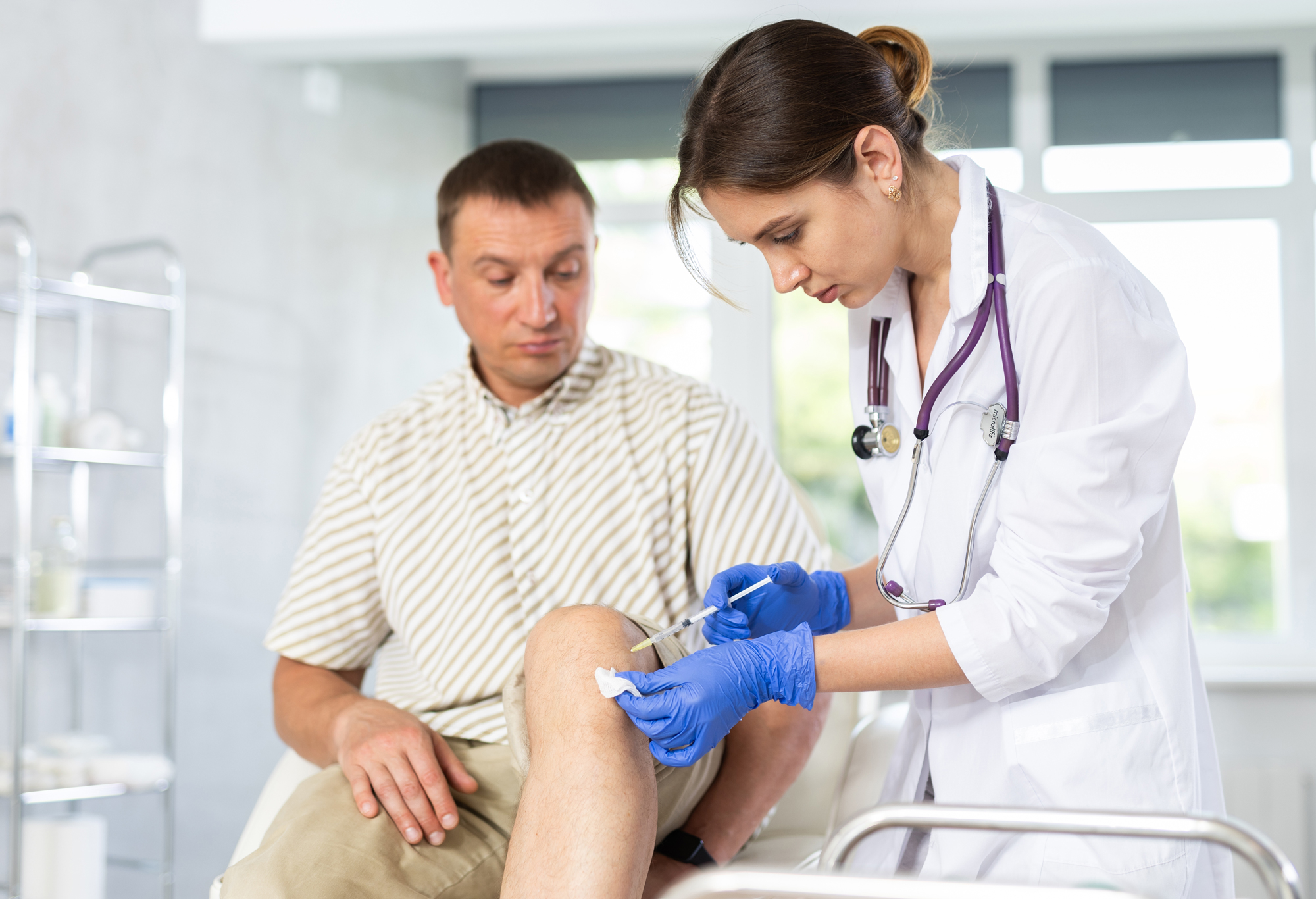 Clinician administering an injection to a seated patient's knee in an exam room